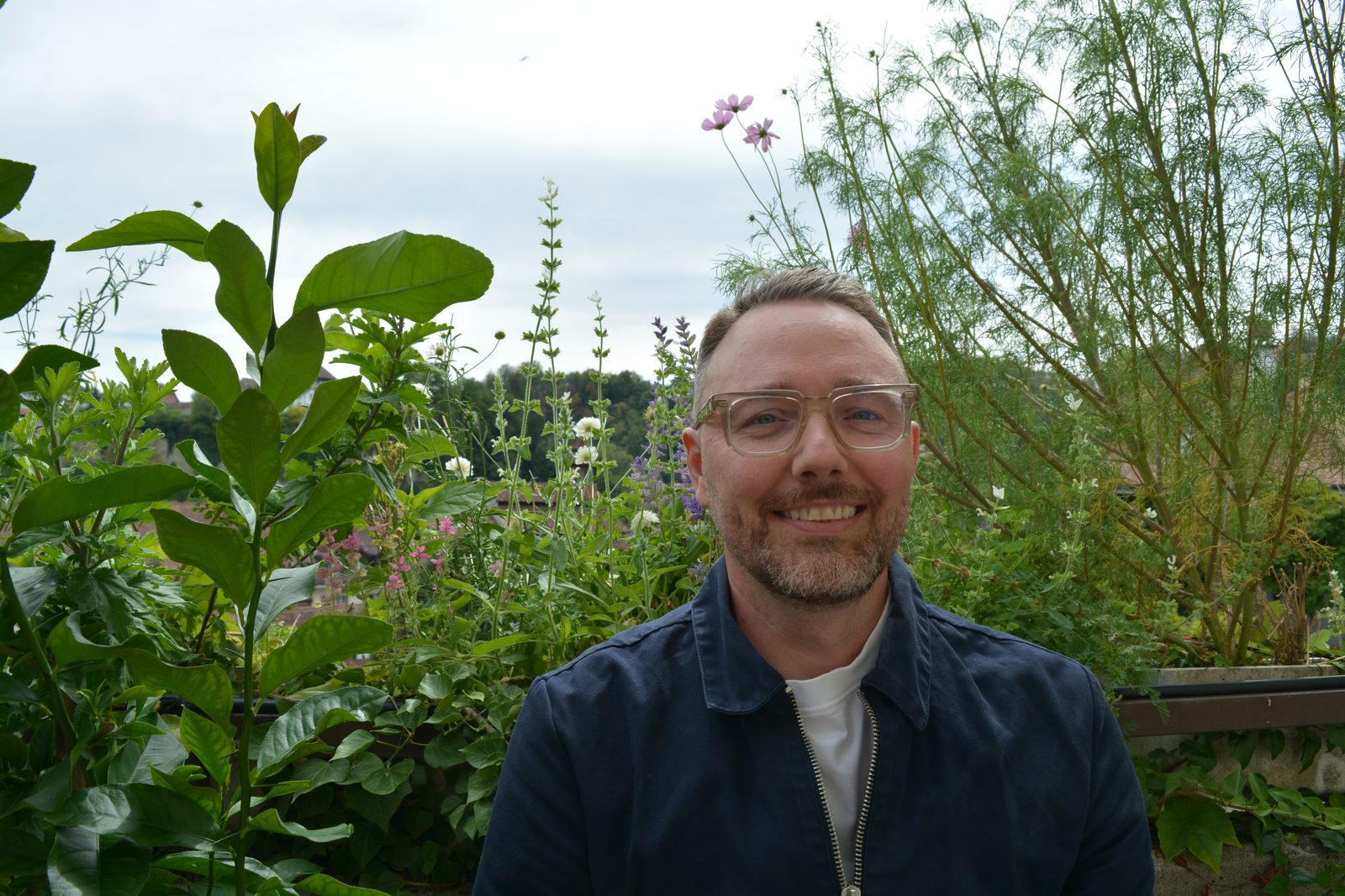 David Ruffieux souriant devant des cosmos et des sauges en fleurs, ciel voilé en arrière-plan